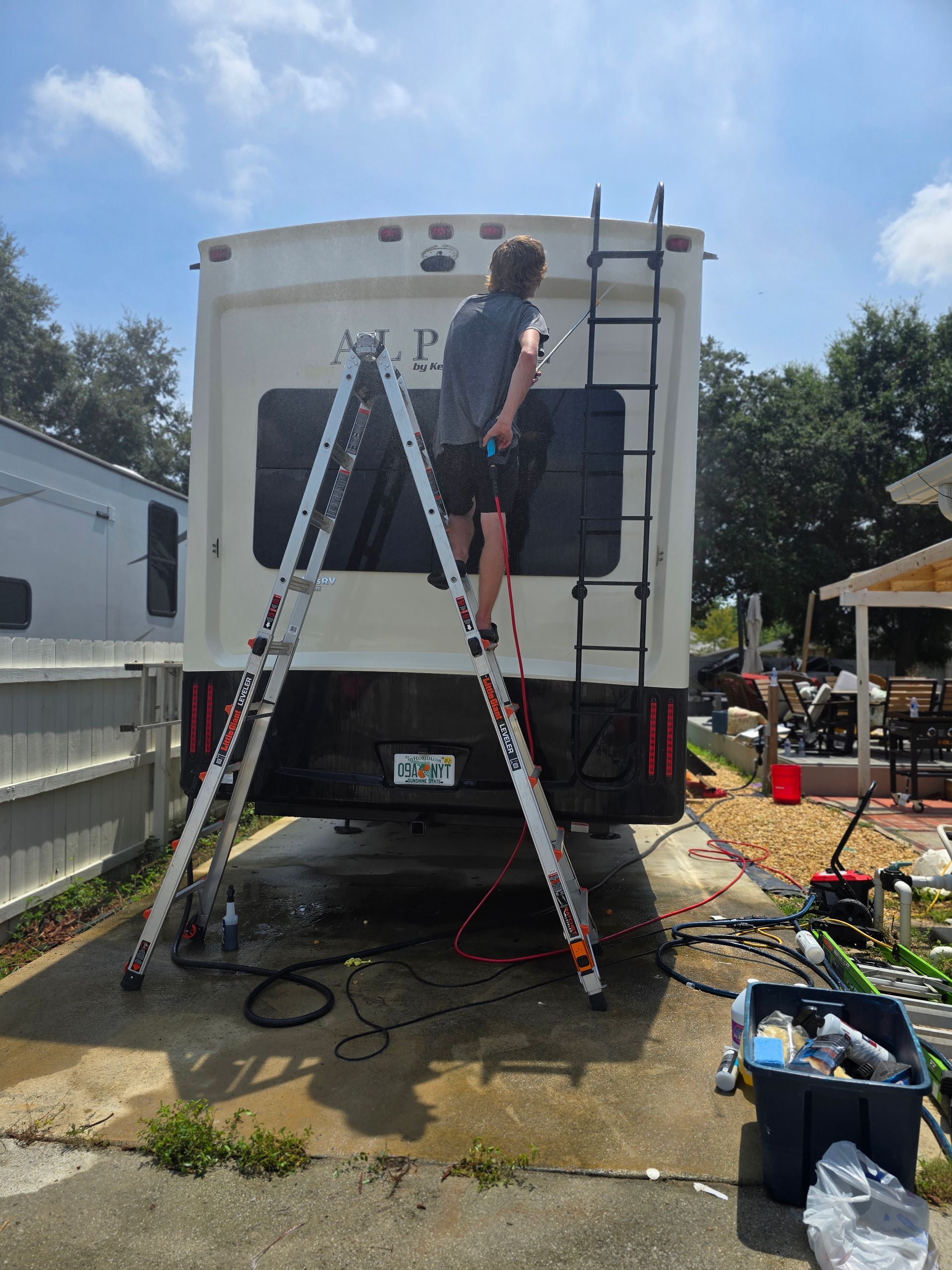 Person on ladder working on rear of RV in a driveway with tools nearby.