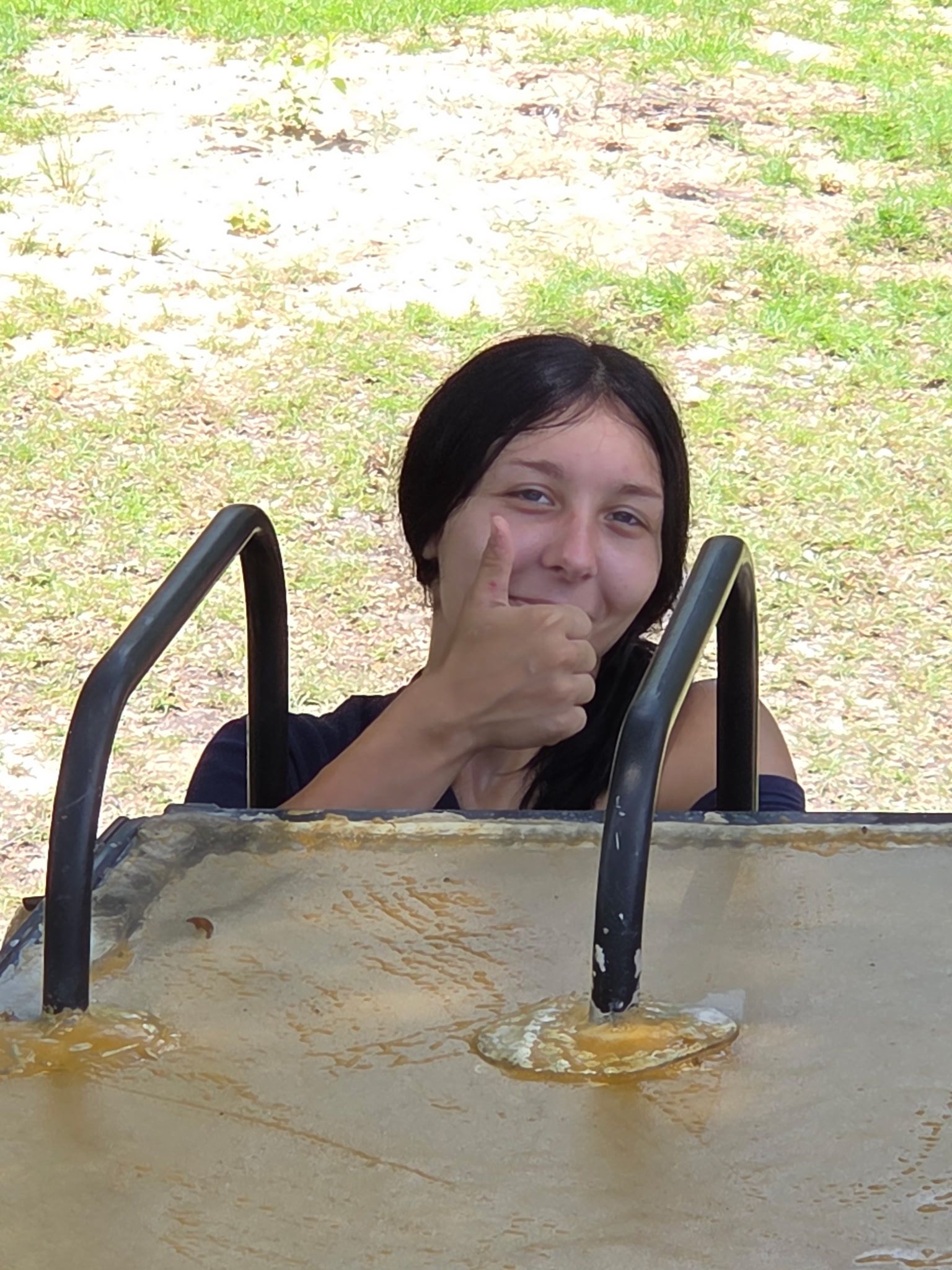 Smiling person giving a thumbs-up while peeking over a playground platform with black rails.