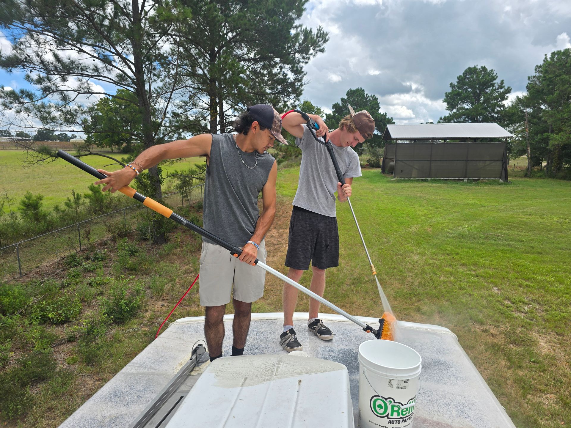 Two people wash a boat on a trailer outdoors, using long-handled brushes and a bucket.