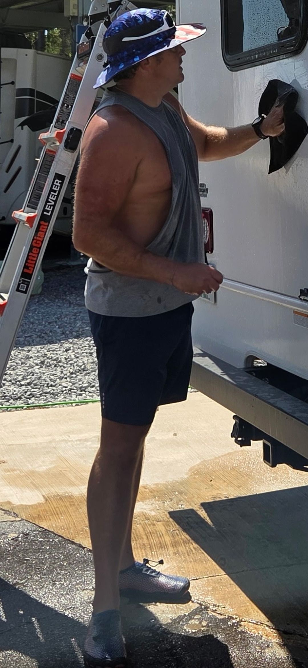 Man standing beside an RV, holding the door and wearing a hat, tank top, shorts, and shoes.