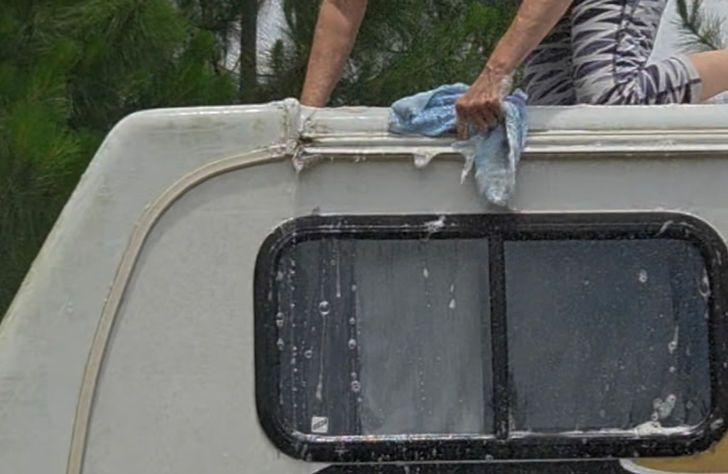 A person wipes a camper window on a gray vehicle outdoors with a blue cloth.