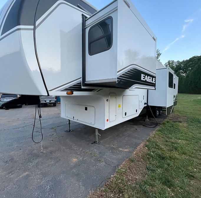 White fifth-wheel RV trailer parked on pavement beside a grassy field under a blue sky