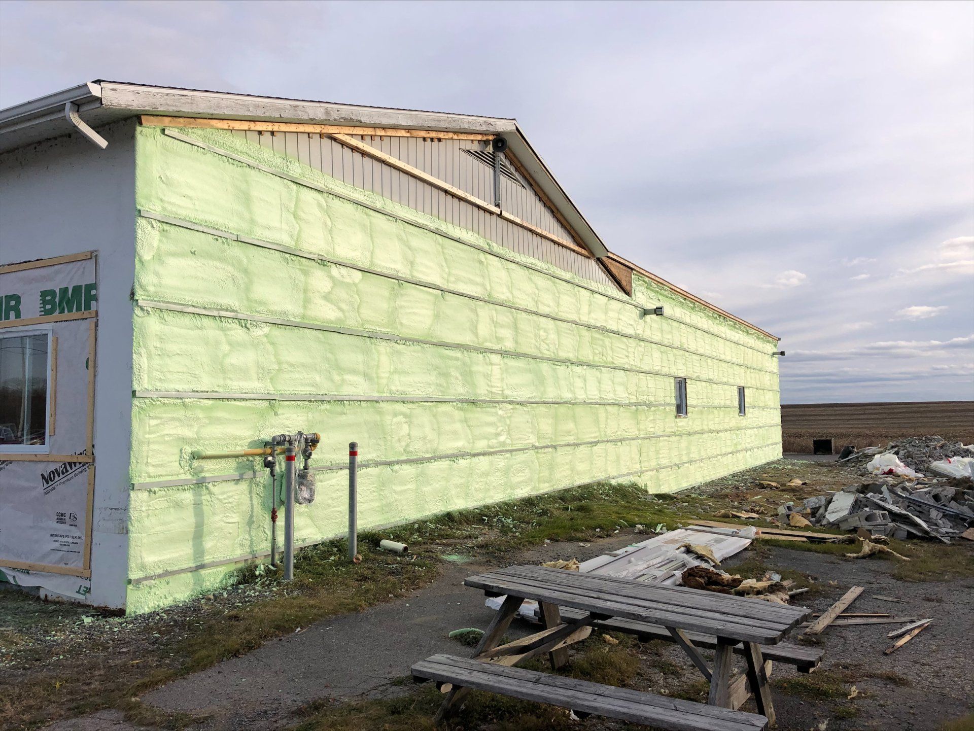 Extérieur du bâtiment avec panneaux isolants verts. Table de pique-nique devant, ciel nuageux.