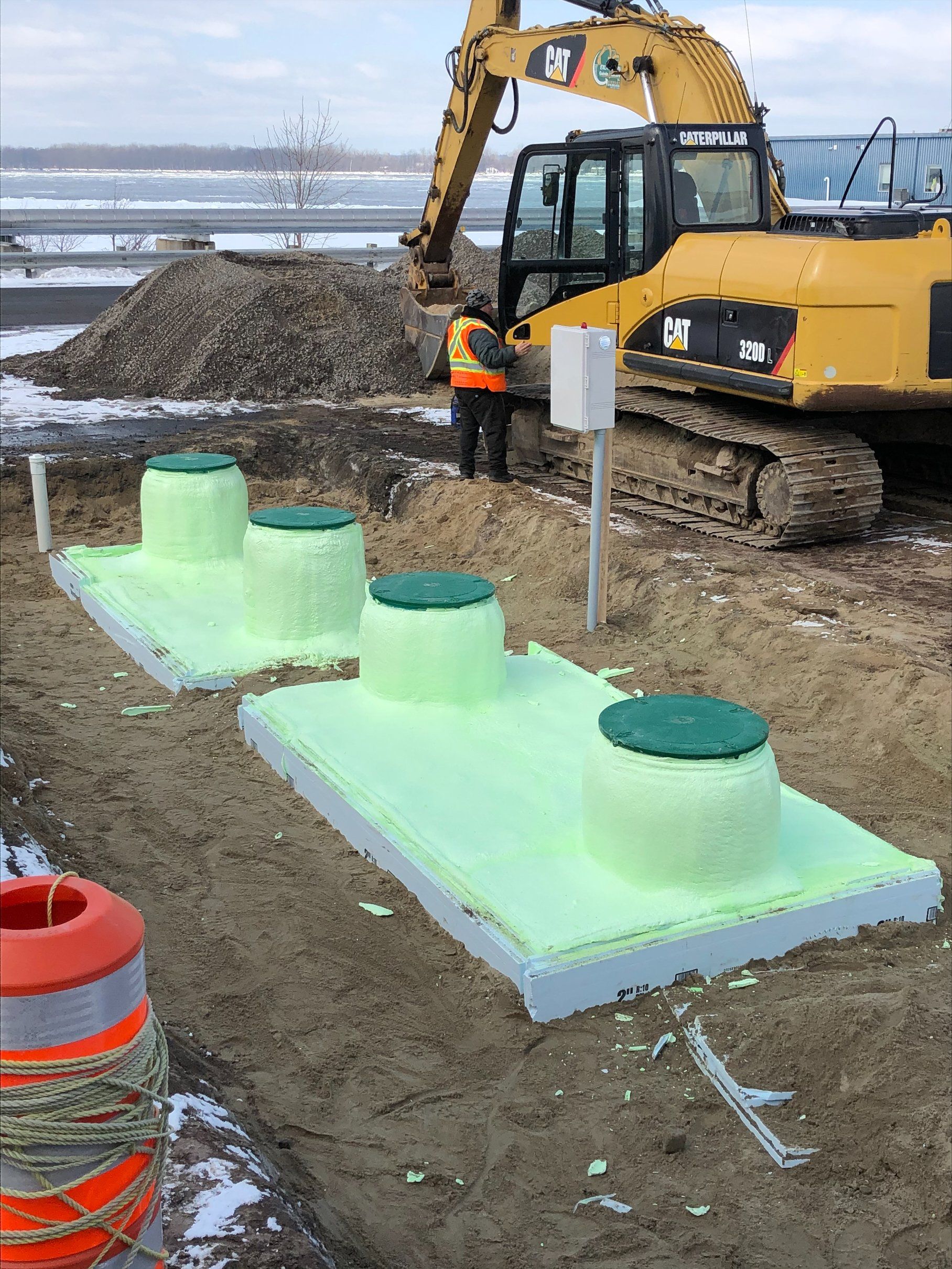 Chantier avec trois fosses septiques en mousse verte. Une pelleteuse jaune et un ouvrier sont visibles à l'arrière-plan.
