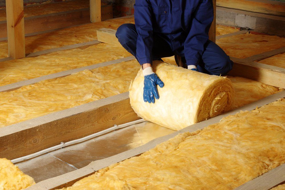 Une personne installe de l'isolant dans un grenier. Des rouleaux d'isolant jaune sont placés entre des poutres en bois.