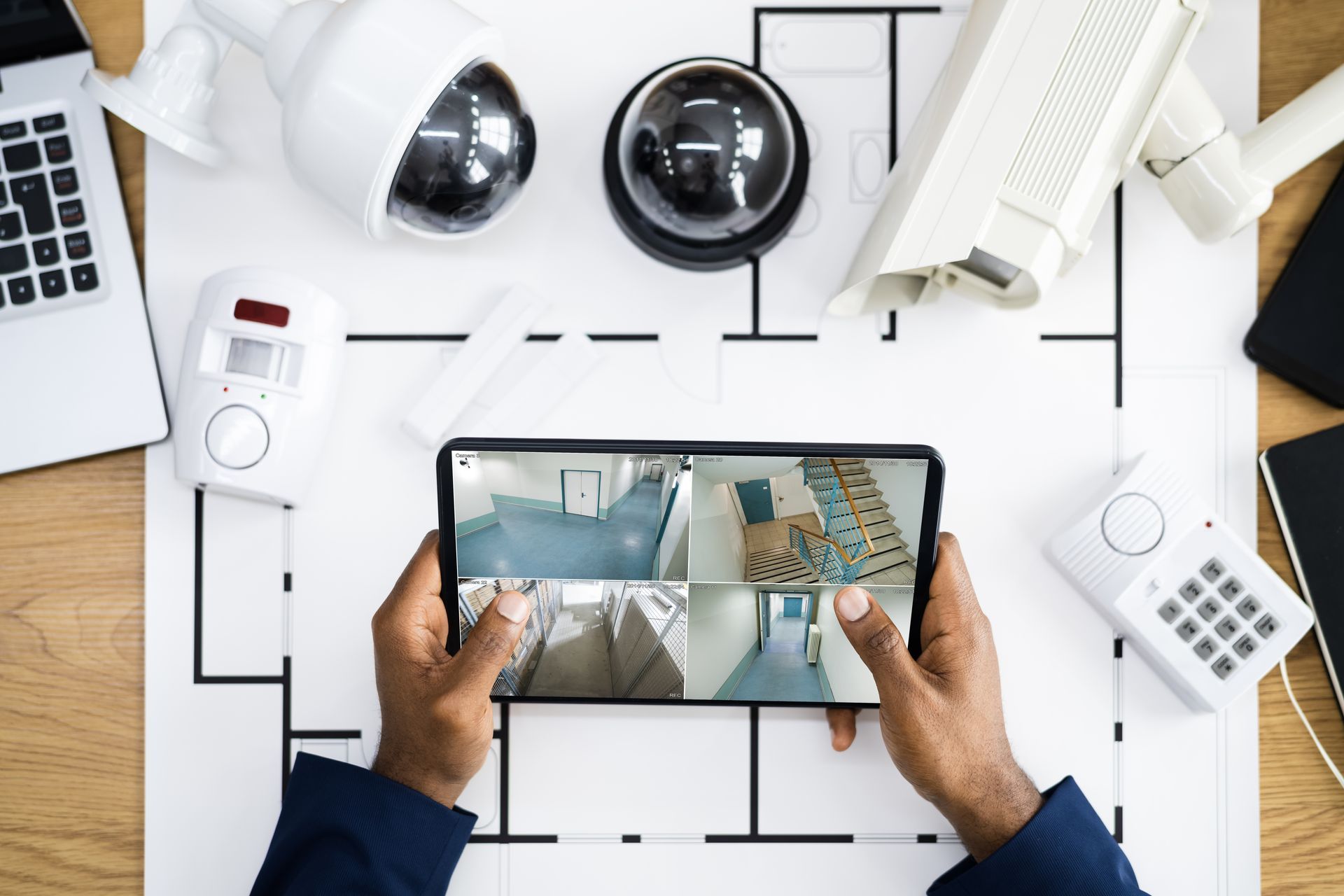 Hands holding a smartphone showing security camera feeds over a home security system layout