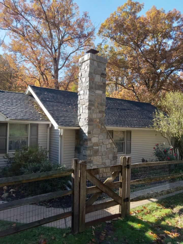 Stone chimney beside a beige house, with a wooden fence and autumn trees in the yard.