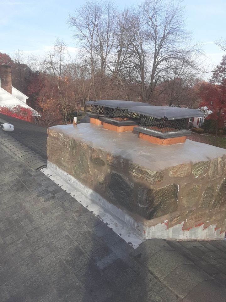 Rooftop chimney and vent with black cap on a shingled roof, trees and sky in the background