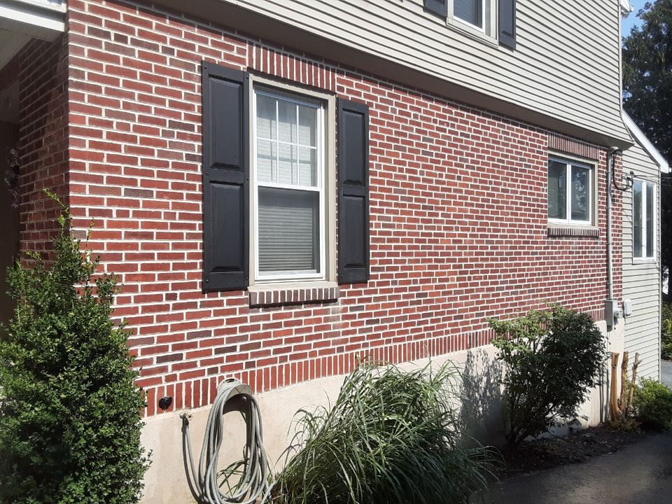 Brick house exterior with black-shuttered window, shrubs, and a garden hose along the foundation