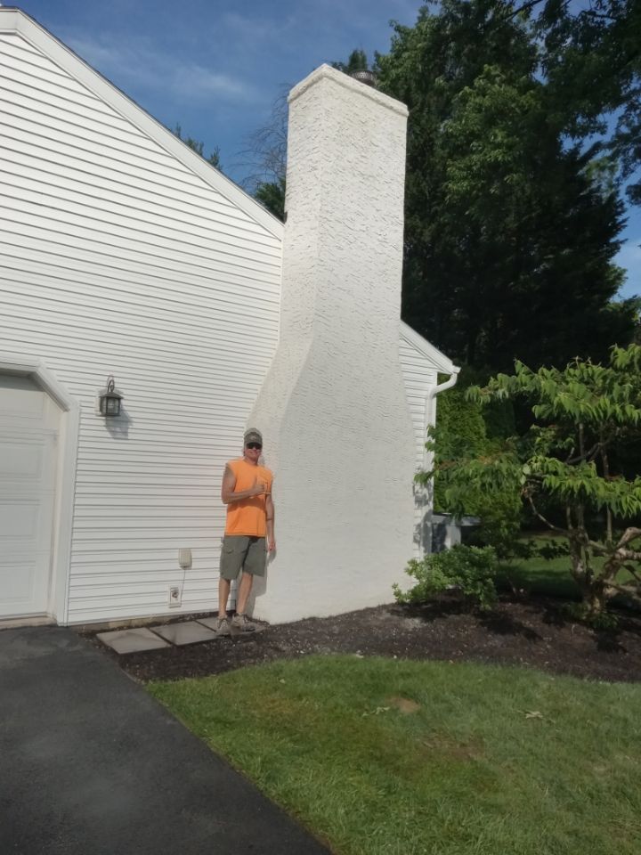 Person in orange shirt standing by a white house chimney and side wall beside a driveway and lawn.