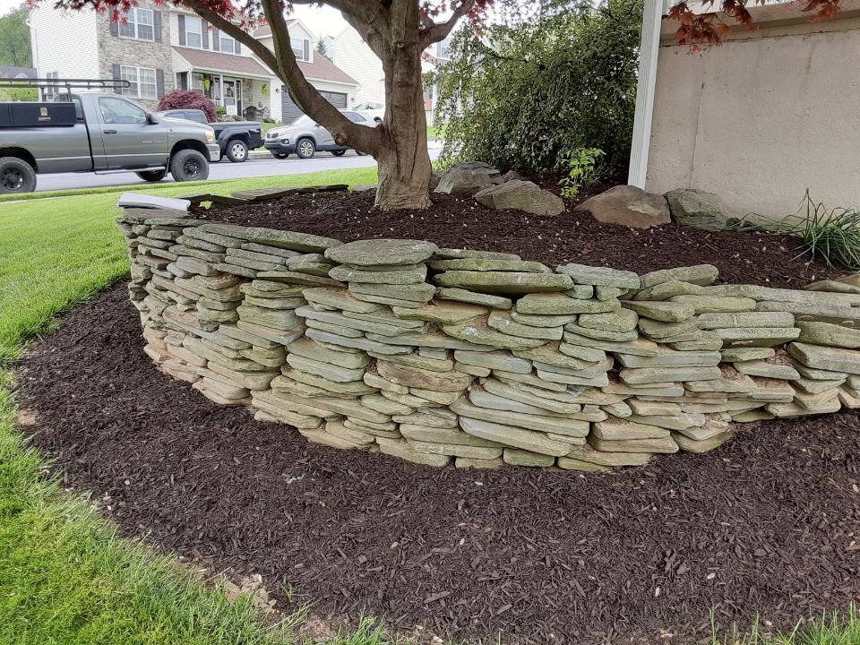 Low stone retaining wall around a landscaped garden bed with mulch and a tree near a street