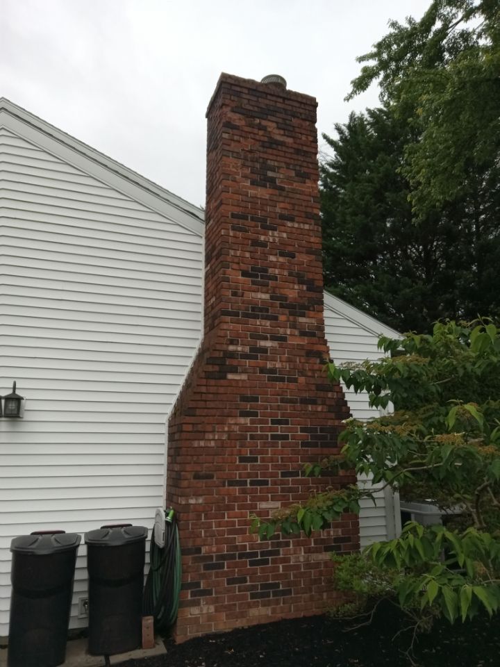 Brick chimney attached to a white-sided house beside black trash bins and greenery