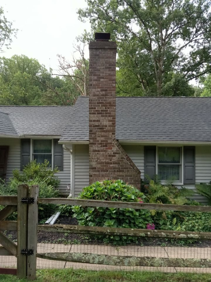 Brick chimney on a single-story house with a gray roof and green shrubs in front.