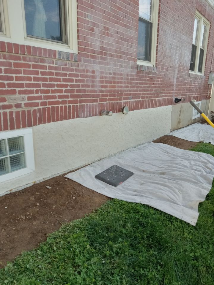 Brick house foundation under repair with white tarp and dirt excavation along the side wall.