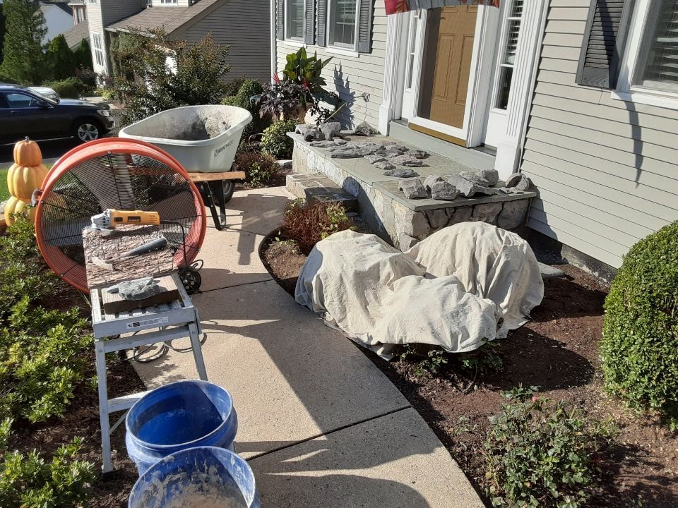 Front yard with covered landscaping stones, wheelbarrow, ladder, and tools beside a house walkway