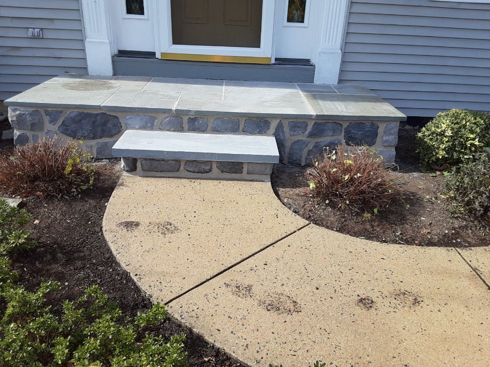 Front porch with stone steps, white bench, and curved concrete walkway beside landscaped shrubs