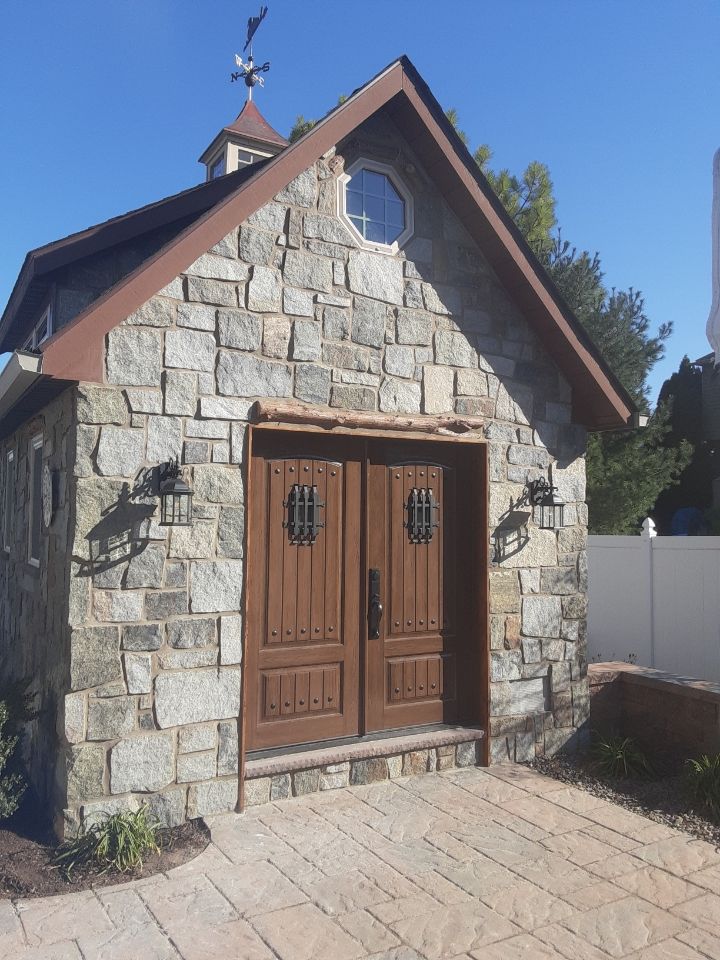 Small stone garden shed with wooden double doors and a pitched roof in a sunny yard.