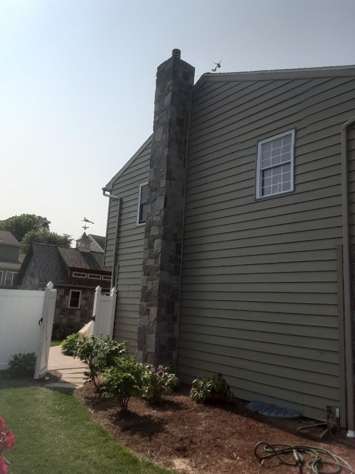 Side view of a gray house with vinyl siding, a stone chimney, and a small landscaped yard.