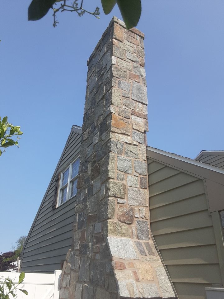 Stone chimney on a house with gray siding and blue sky.