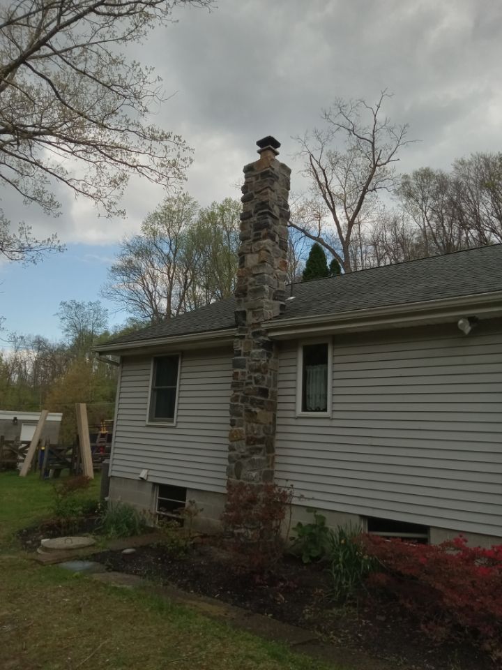 White house with a stone chimney and gray siding, viewed from the yard under a cloudy sky