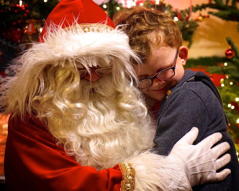 Boy hugging Santa Claus, red suit, white beard, Christmas tree in background.
