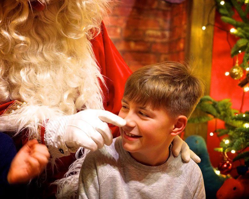Santa playfully touches a boy's nose. Boy smiles. Red and green holiday decor in the background.
