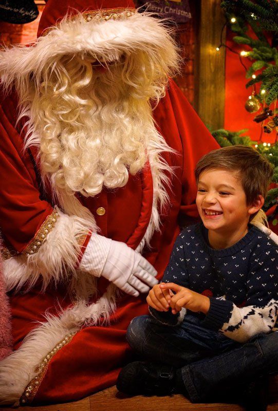 Boy laughs with Santa Claus, sitting next to a Christmas tree.