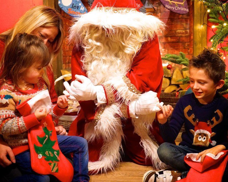 Santa with two children and a woman, looking at stockings, in a decorated room.
