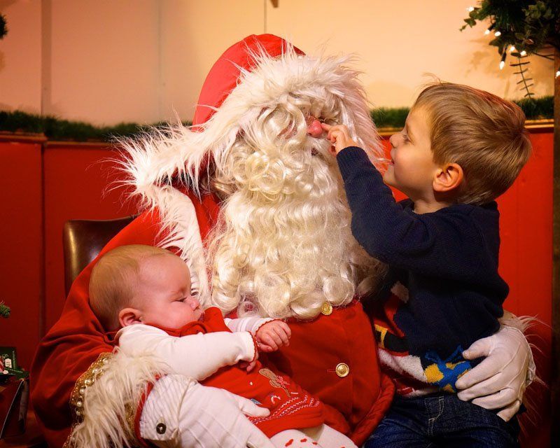 Santa Claus with a baby and a toddler, the toddler touching Santa's nose. Red and white outfits.
