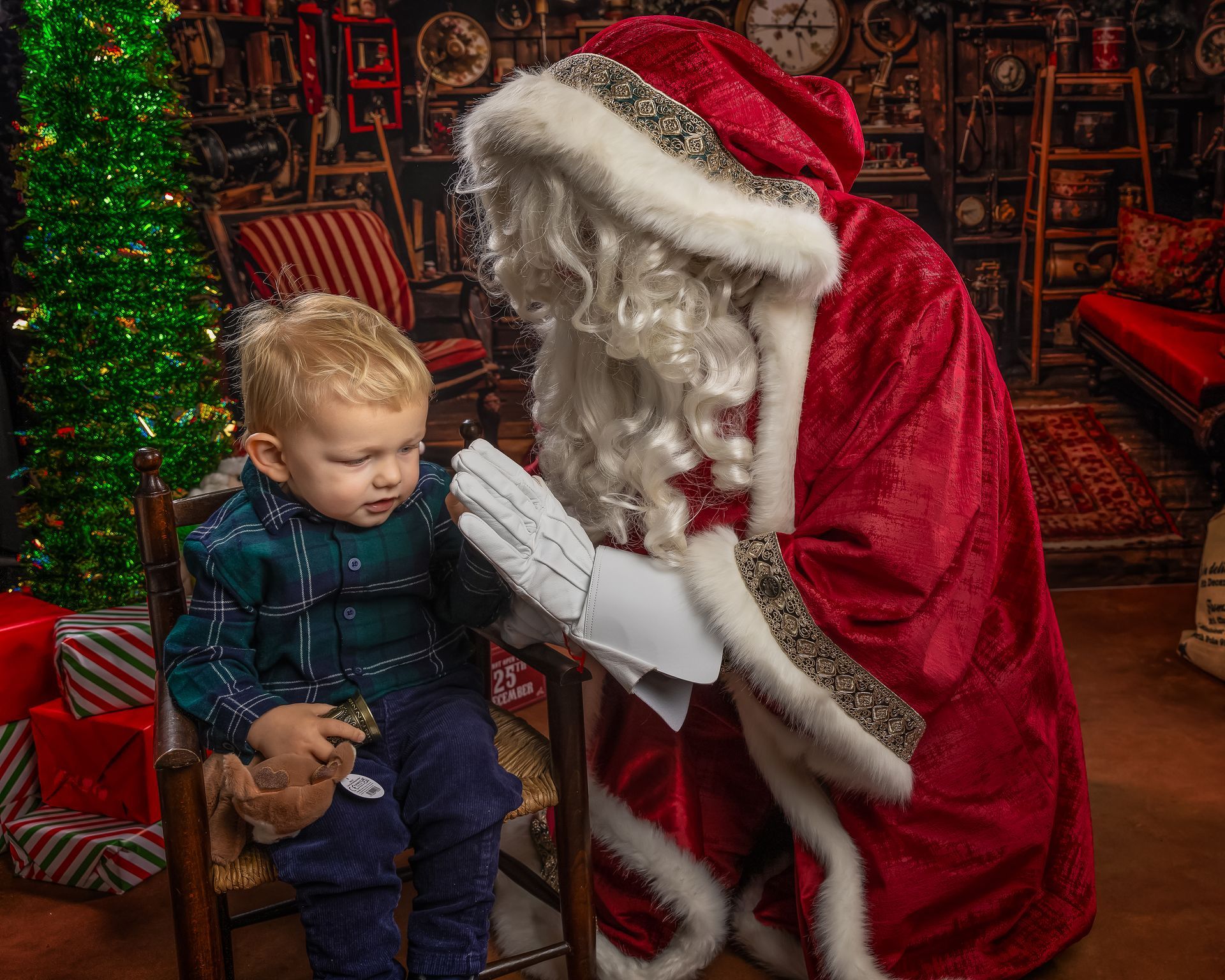 Santa playfully touches a boy's nose. Boy smiles. Red and green holiday decor in the background.