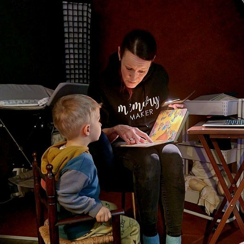 Woman reading a book to a young child. Both are indoors; the child is seated, listening intently.