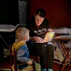 Woman reading a book to a young child. Both are indoors; the child is seated, listening intently.