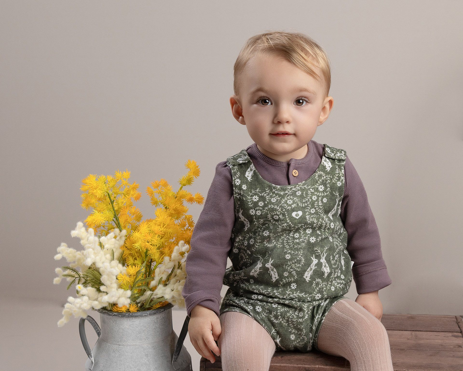 A toddler sits on a wooden surface, wearing a green outfit and purple long-sleeved shirt, with flowers in a metal pitcher.