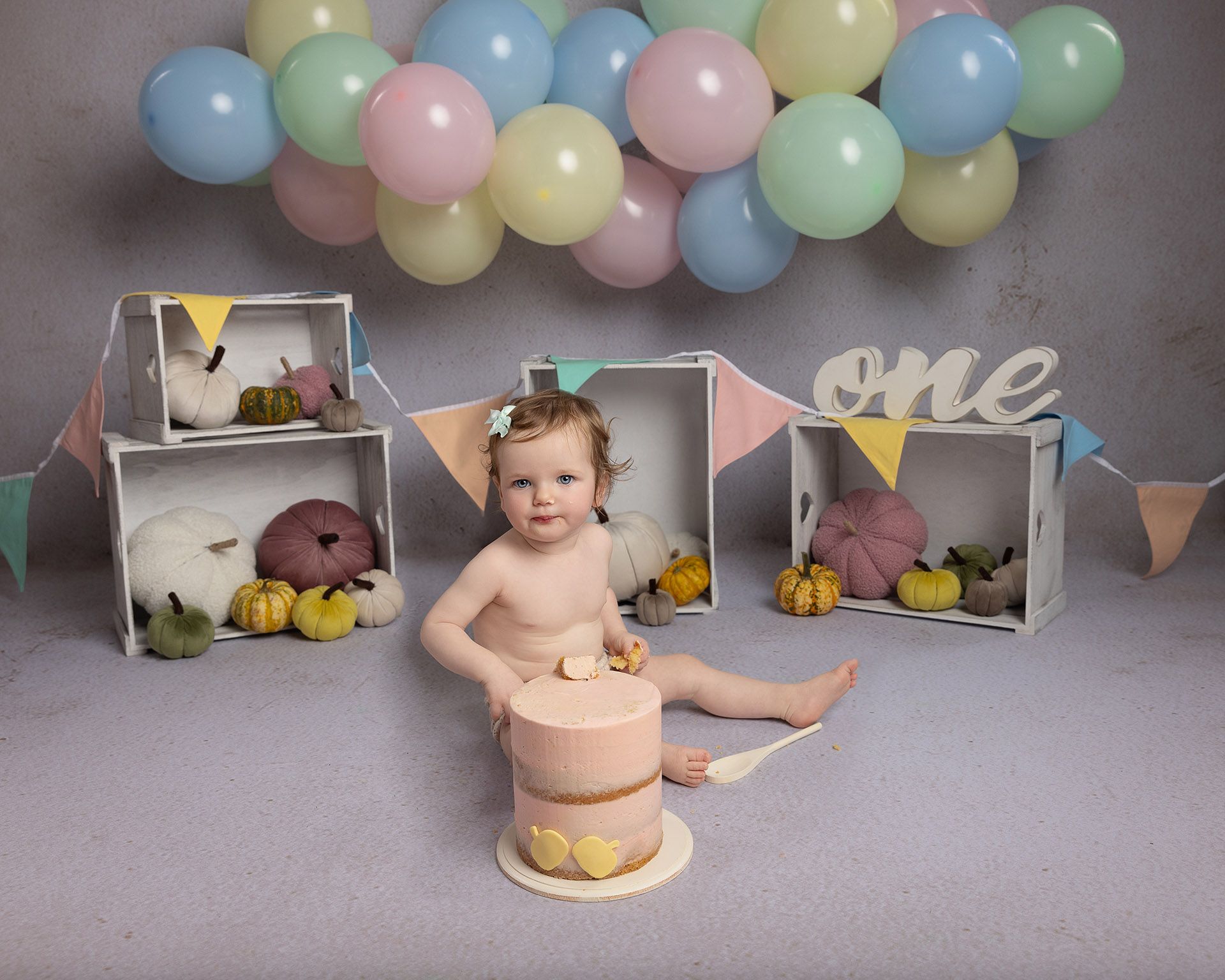 Baby celebrating first birthday, smeared with cake, surrounded by balloons and pumpkins.