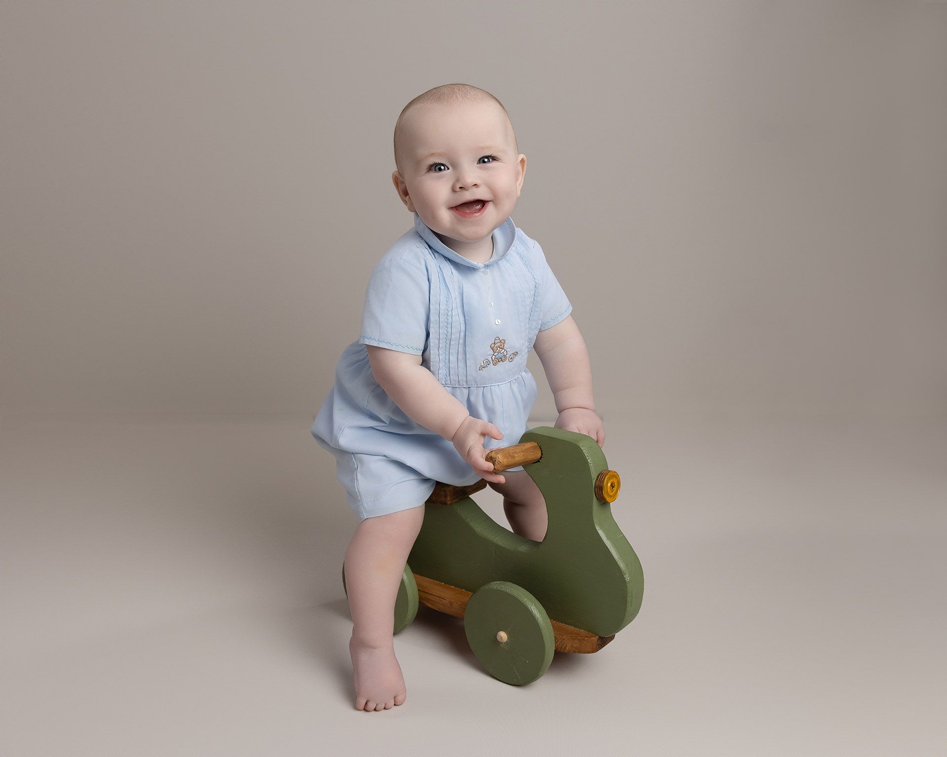 Smiling baby in blue outfit on a green wooden toy, light background.