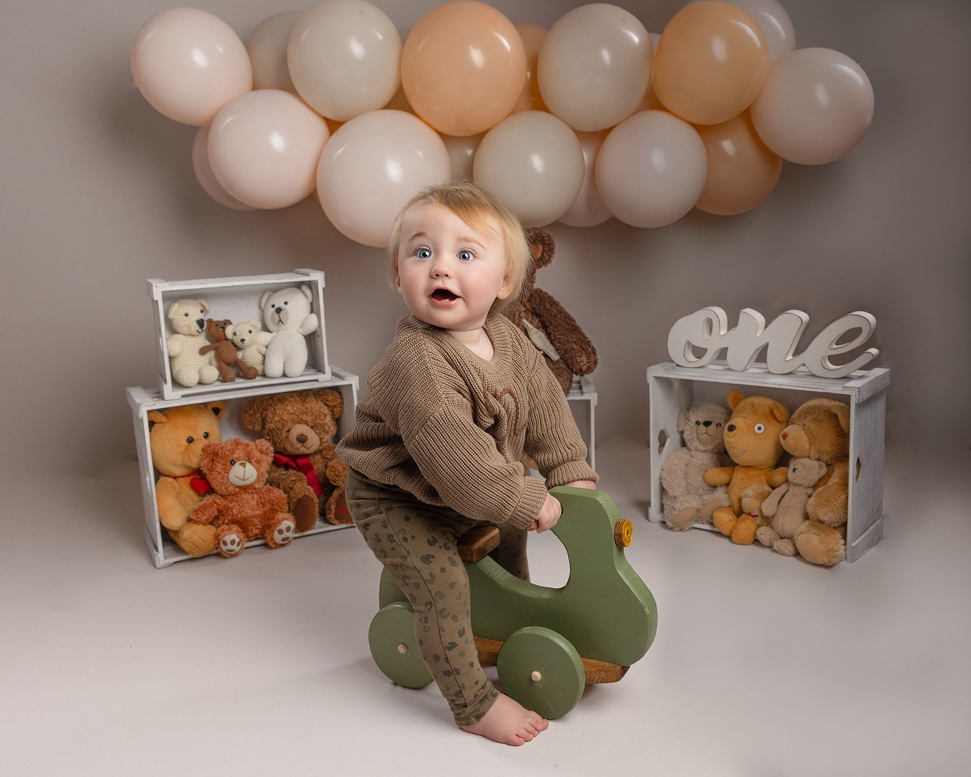 A baby celebrating their first birthday, riding a green toy car in a room decorated with teddy bears and balloons.