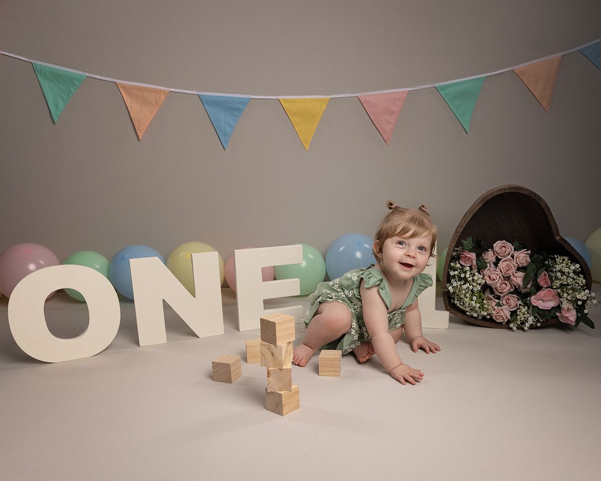 A baby girl smiles, crawling toward camera, surrounded by balloons,