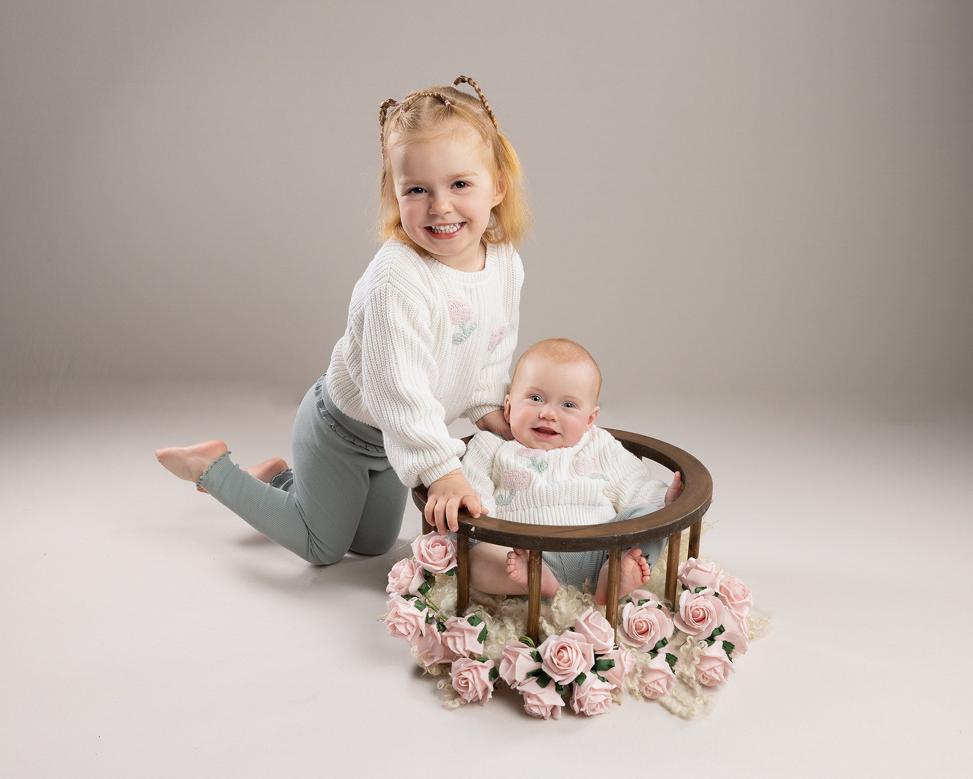 Girl kneels by baby in a circular bed with pink roses. Both wear white sweaters, smiling against a grey background.