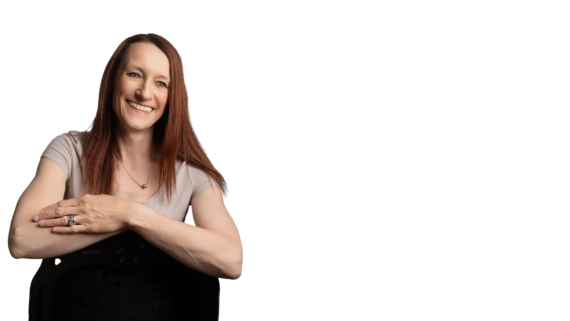 Woman with auburn hair smiles, leaning on a chair, arms crossed, wearing a light colored shirt.