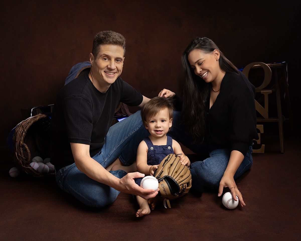 Family of three, wearing black and jeans, smiles for the camera, baby in the center holding a baseball glove, brown background.