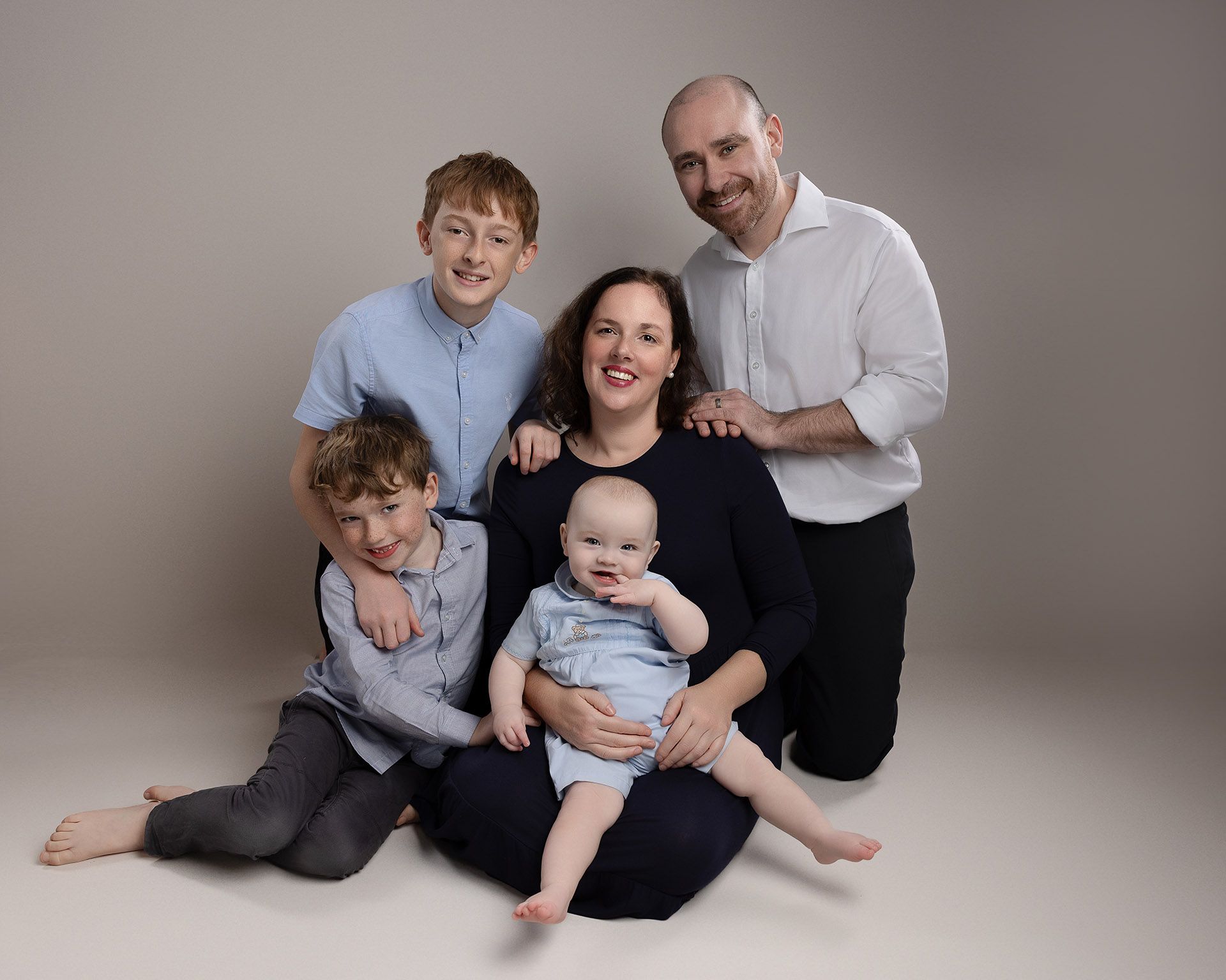 Family portrait: mom holding baby, two sons flanking her, dad kneels behind. Neutral background, smiling faces.