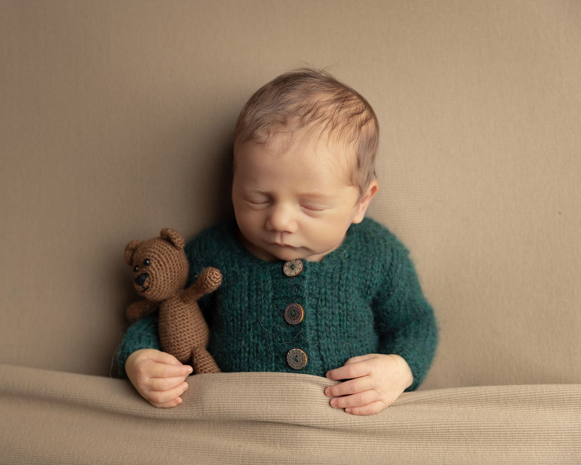 Newborn baby sleeping in green sweater, holding a teddy bear.