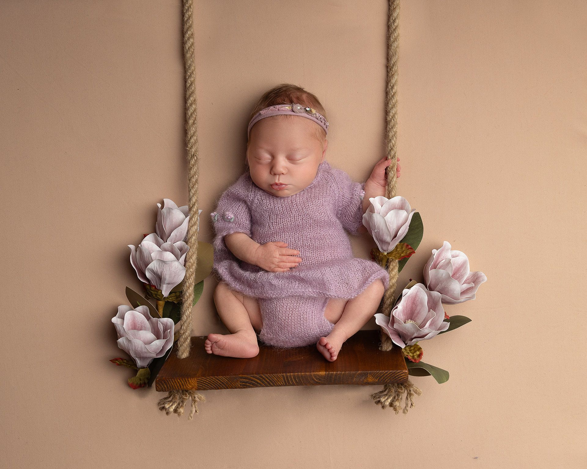 Newborn baby asleep on a wooden swing, wearing a lilac outfit and headband, surrounded by flowers.
