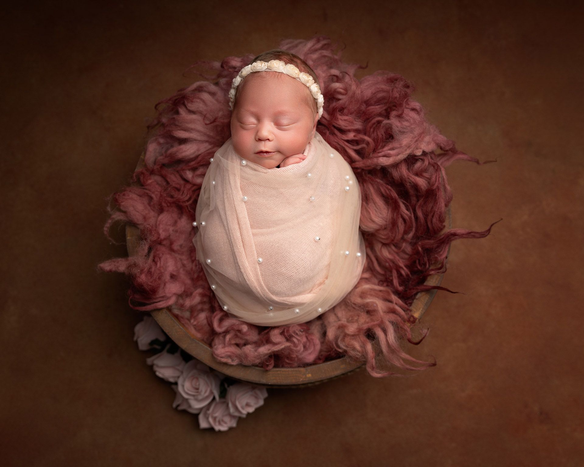 Newborn baby wrapped in a pink blanket with a pearl design, in a bowl with pink fur, wearing a headband.