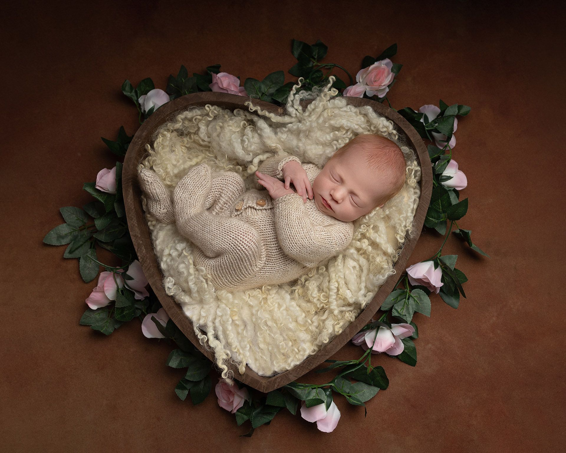 Newborn baby sleeping peacefully in heart-shaped bed of soft, cream-colored fur surrounded by flowers.