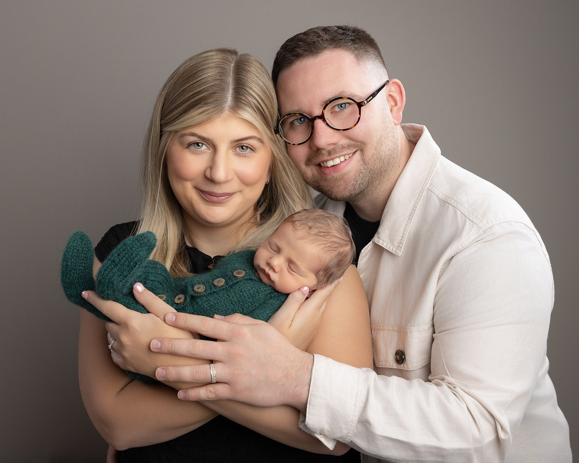 New parents smiling, holding newborn baby in green outfit, against a gray backdrop.