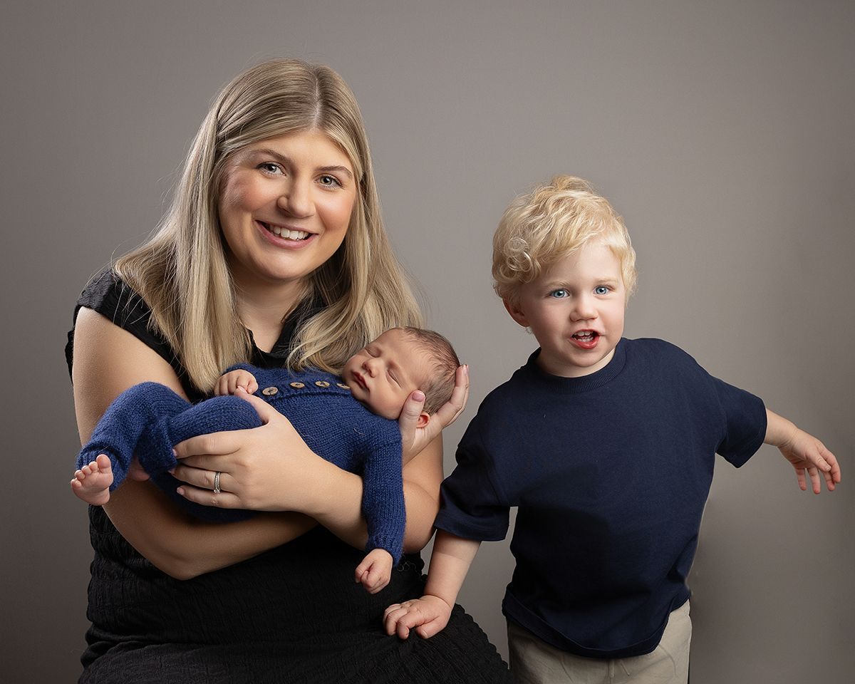 Woman holding newborn, toddler beside her. They are smiling, against a gray background.