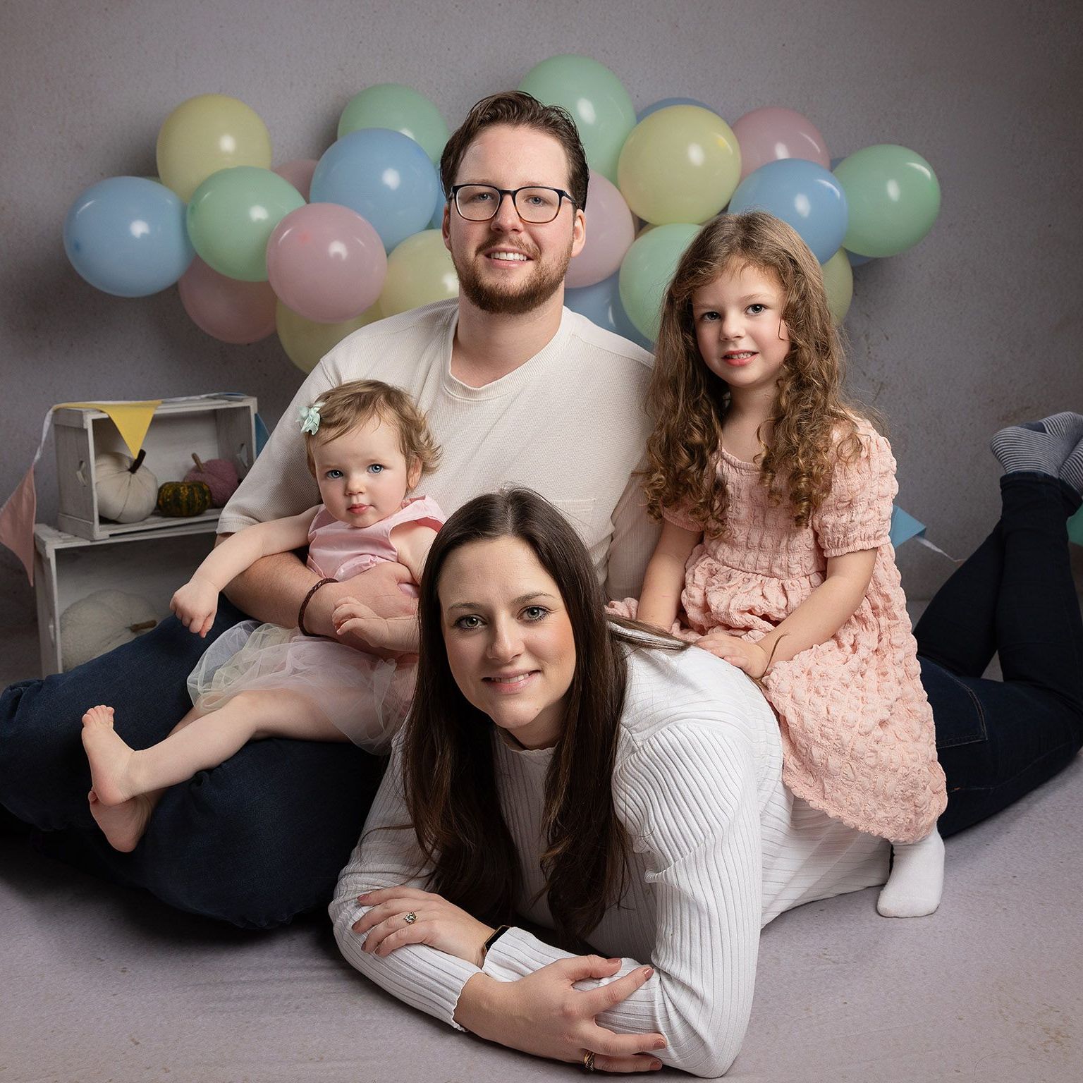 Family of four smiling, poses with balloons. Man and one girl sit, woman lies down. Soft colors.