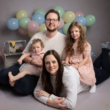 Family of four smiling, poses with balloons. Man and one girl sit, woman lies down. Soft colors.