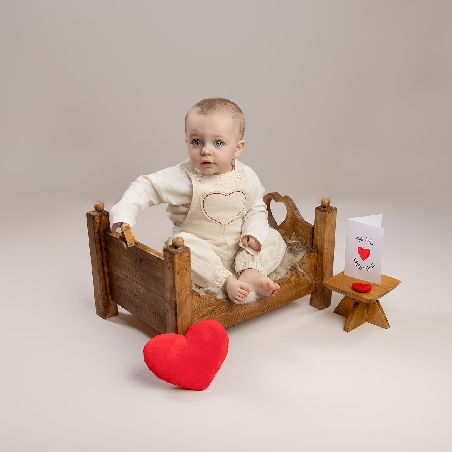 Baby in cream outfit sits in wooden bed, looking left, with red heart and card nearby.